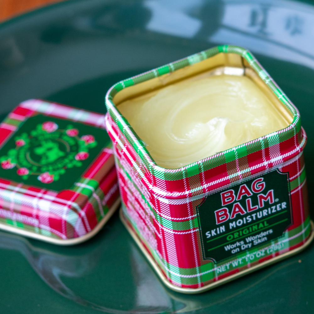 Open tin of Bag Balm skin moisturizer with plaid design on a reflective surface